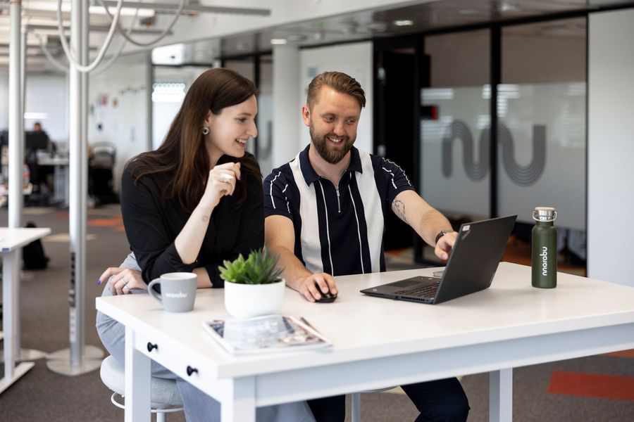 The image shows two people collaborating at a desk in a modern office setting. They are both smiling and engaged with a laptop, which the man is pointing at. The woman holds a pen and appears attentive. The desk has a small potted plant, a coffee mug, a reusable water bottle, and a magazine. The environment is open and bright, with glass walls and visible workstations in the background. The branding on the items ("marabu") suggests a shared company or team identity.