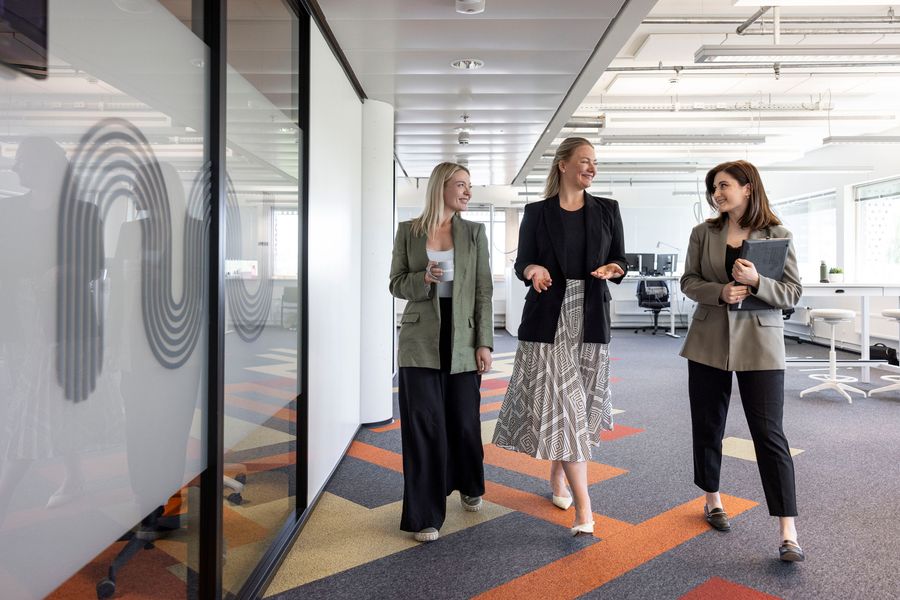 The image shows three professionally dressed women walking together in a modern, open-plan office. They are engaged in conversation and appear relaxed and cheerful. One is holding a cup, another has her hands gesturing while speaking, and the third is carrying a folder. The office space features bright lighting, contemporary furniture, and colorful patterned carpet tiles. A glass wall with a wavy logo design is visible on the left side of the image.