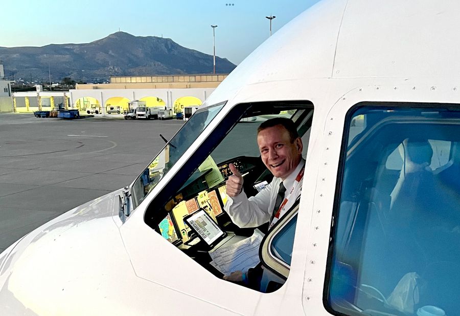 The image shows a smiling pilot sitting in the cockpit of an airplane, giving a thumbs-up gesture. The aircraft is parked on the tarmac, and various airport service vehicles can be seen in the background along with a mountainous landscape. The scene is brightly lit, possibly during early morning or evening, with a clear sky above.