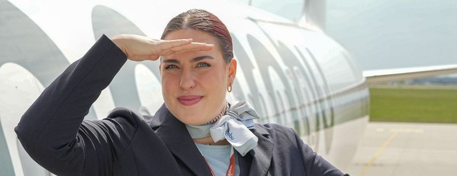 The image shows a flight attendant standing outside an airplane near the boarding stairs, smiling and holding one hand to her forehead as if shielding her eyes from the sun. She is dressed in a professional navy-blue uniform with a scarf and lanyard displaying a name badge. The aircraft behind her displays part of the airline's logo or branding along the fuselage, and the scene appears to be taken on a clear day at an airport apron.